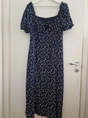 Marinblå blommig långklänning - Söt marinblå långklänning från Gina Tricot med vitt blommönster, puffiga korta ärmar och knyt framtill vid urringningen. Klänningen har en avslappnad passform och är perfekt för sommarens alla tillfällen. Elastisk. Perfekt, som ny. Storlek XL
