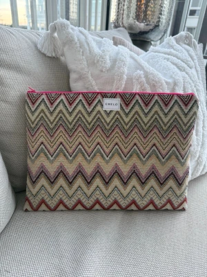 Zigzag-mönstrad datorväska från Emelo - Säljer en snygg datorväska från Emelo med färgglatt zigzag-detaljer i beige, rött, rosa och svart. Väskan har en rektangulär form och stängs med en rosa dragkedja. Materialet är textil med ett mjukt innerfoder. Perfekt för att bära din laptop med stil. Passar laptop med maxmått: 33x24cm
