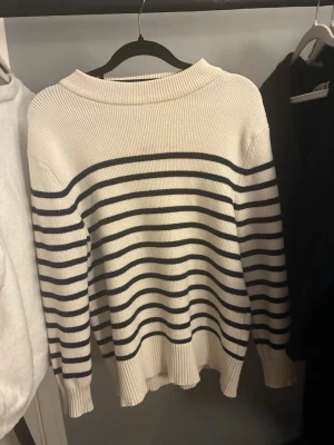 Randig stickad tröja från Zara - Säljer en krämvit stickad tröja från Zara med marinblå horisontella ränder på ärmarna och över bröstet. Tröjan har rund halsringning och ribbstickade muddar. Perfekt för dig som gillar klassisk och stilren design.❤️