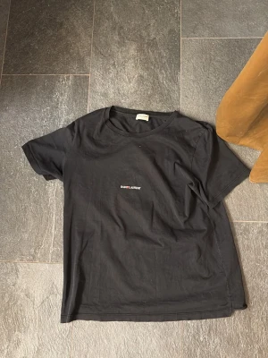 Svart t-shirt från Saint Laurent - Svart t-shirt från Saint Laurent med liten vit och röd logga på bröstet. Klassisk rund halsringning och korta ärmar. Tillverkad i mjuk bomull för en skön känsla och avslappnad passform. 