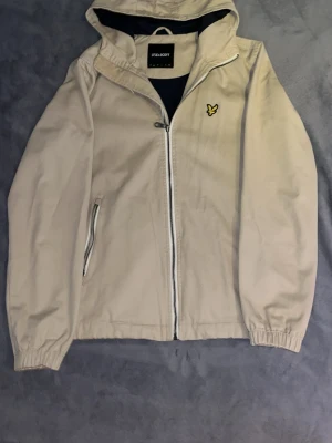 Beige vindjacka från Lyle & Scott - Stilren beige vindjacka från Lyle & Scott med huva och dragkedja framtill. Jackan har ett broderat gult logomärke på bröstet och två fickor med dragkedja. Perfekt för blåsiga dagar och enkel att matcha med olika outfits.