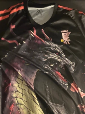 Japan JFA svart draktröja - Cool svart sporttröja från Japan JFA med en stor, detaljerad drake tryckt över hela framsidan. Tröjan har röda detaljer på axlarna och JFA-logga på bröstet. Perfekt för dig som gillar unika och snygga sportkläder.