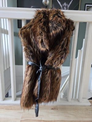 Brun fuskpälsväst med bälte - Säljer en snygg brun fuskpälsväst med svart knytband i midjan i Nyskick. Västen har en fluffig och mjuk känsla med naturliga färgskiftningar i pälsen. Den har fickor samt hakar till knäppe. Storlek 44, men skulle säga att den passar mellan 40-44 beroende på vad man har under.