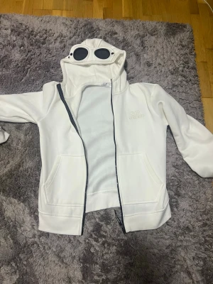 Vit hoodie med goggles från C.P. Company - Säljer en vit hoodie från C.P. Company med hel dragkedja och ikoniska goggles i huvan. Hoodien har två fickor framtill och är tillverkad i mjukt material, perfekt för en cool och unik streetwear-look.