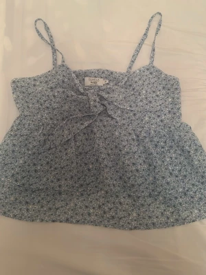 Blommig blå linnetopp från Pamela/Nakd - Supersöt blå linnetopp från Pamela med små blommor och rosett framtill. Tunt och luftigt material, perfekt för varma dagar.