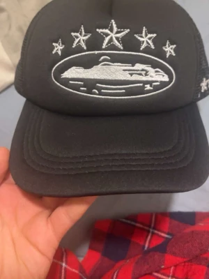 Corteiz keps - Snygg svart truckerkeps med vit brodyr framtill som visar ett fartyg och fem stjärnor ovanför. På sidan finns fler broderade stjärnor. Kepsen har mesh på sidorna och bak för extra ventilation och justerbar passform. Perfekt för dig som gillar unik och cool stil. Helt ny btw.