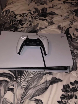 PlayStation 5 med DualSense-kontroll - PlayStation 5 spelkonsol i vit och svart design med tillhörande DualSense-kontroll. Konsolen har en futuristisk, rektangulär form och är tillverkad i plast. Perfekt för gaming och underhållning.