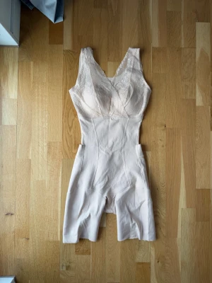 Beige shapewear med spetsdetaljer - Säljer en beige shapewear-body med breda axelband och v-ringning. Överdelen har fina spetsdetaljer och kupor, medan nederdelen är slät och går ner till låren. Perfekt för att forma och ge stöd under kläder. Materialet är stretchigt och bekvämt.
