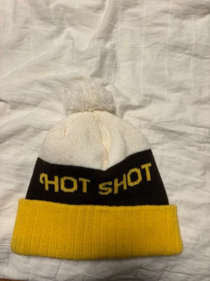 Gul och vit mössa med text - Snygg mössa med vit topp och tofs, svart mittparti med gul text 'HOT SHOT' och bred gul uppvikt kant. Perfekt till afterski 🫰🏼