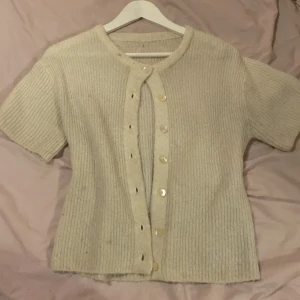 Beige gina tricot stickad kofta med kort ärm  - Snygg beige ribbstickad kofta med korta ärmar och rund halsringning. Koftan har knäppning framtill med pärlemorknappar och en mjuk, mysig känsla. Perfekt att slänga över en topp för en chill look.