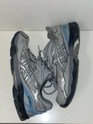 Gråa asics  - Säljer ett par gråa Asics sneakers med blå och svarta detaljer. Skorna har en chunky sula och klassisk mesh- och syntetöverdel med Asics logga på sidan. Perfekta för dig som gillar sportig och retro stil. OBS SÖNDER INUTI HÄLEN 