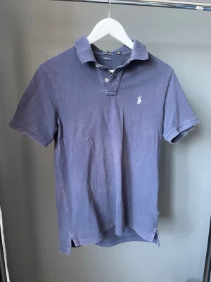 Marinblå pikétröja från Polo Ralph Lauren - Säljer en klassisk marinblå pikétröja från Polo Ralph Lauren med vit logga på bröstet. Tröjan har korta ärmar, krage och knappar framtill. Perfekt för dig som gillar en stilren och sportig look.