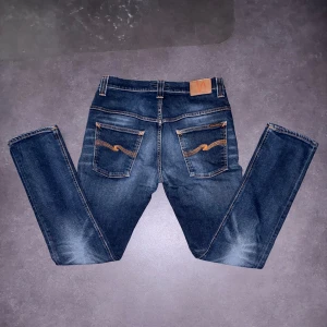 Blå jeans från Nudie Jeans Co 32W 32L - Säljer ett par snygga blå jeans från Nudie Jeans Co 42cm midja och 108cm längd med klassisk femficksdesign och coola orangea sömmar på bakfickorna. Jeansen är tillverkade i 100% ekologisk bomull och har en straight passform med normal midja. Perfekt för dig som gillar stilrena och tidlösa jeans.