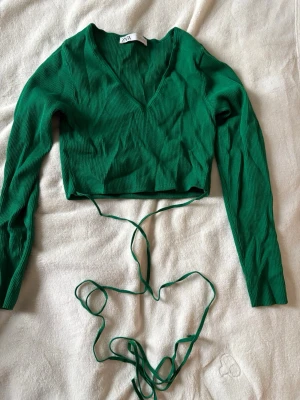 Grön croppad topp från Zara - Trendig grön croppad topp från Zara med långa ärmar och djup v-ringning. Toppen är ribbad och har långa knytband i midjan som ger en snygg detalj i storlek L