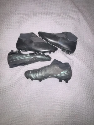 Nike Mercurial svarta fotbollsskor - Säljer ett par svarta Nike Mercurial fotbollsskor med strumpliknande skaft och dobbar för spel på gräs. Skorna har en modern, strömlinjeformad design och texturerad ovandel för bättre bollkontroll. Snygg diskret logga på sidan och lätta material för snabbhet. Paret högst upp är i strl 43,5 och paret underst är i 42. 