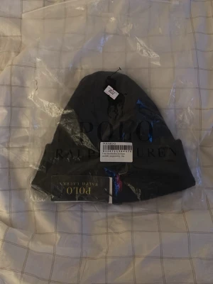 Svart Polo Ralph Lauren mössa - Snygg svart Polo Ralph Lauren mössa med klassisk röd logga. Mössan har uppvikt kant och är tillverkad i mjukt material som håller dig varm. Perfekt accessoar för kalla dagar och stilren look.