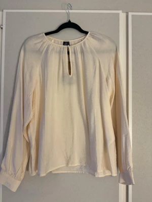 Krämvit blus  - Stilren krämvit blus från Vero Moda med långärmad design och avslappnad passform. Blusen har en rund halsringning med nyckelhålsöppning framtill och knäppning. Tillverkad i ett lätt, glansigt material som ger en mjuk känsla. 