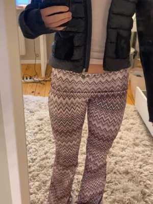 Lila mönstrade bootcut leggings - Säljer ett par lila och vita leggings med zickzack-mönster och bootcut-modell. De har en bred, vikt linning och sitter tajt upptill med utsvängda ben. Perfekta för dig som gillar retrostil och vill sticka ut.
