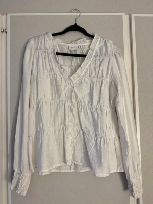 Vit blus  - Söt vit blus från Vila med volangkant längs v-ringningen och framtill. Blusen har långärmade ärmar med smock vid ärmsluten och ett lätt rynkat tyg som ger en avslappnad och trendig look. (En av dem man sätter fast knappen i har lossnat på ena sidan - se bild 3, inget man tänker på dock) 