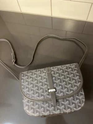 Grå axelväska  - Snygg grå axelväska  med klassiskt mönster och justerbar axelrem. Väskan har lock med spänne och detaljer i läder. Perfekt för dig som gillar stilrena accessoarer med lyxig känsla. Skickas inom 24 timmar