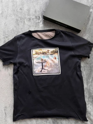 Svart T-shirt med tryck från Limitato - Svart T-shirt från Limitato med ett stort färgglatt tryck framtill som föreställer en kvinna vid en pool. Klassisk rund halsringning och korta ärmar. Perfekt för dig som vill sticka ut med en unik design.
