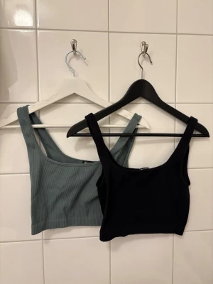 Ribbad crop top från Zara, 2-pack - Två ribbade crop tops från Zara, en i svart och en i gråblå nyans. Båda är ärmlösa med breda axelband och har en tight passform. Perfekta att styla med jeans eller kjol för en trendig look.