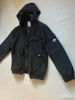 Svart vindjacka från M - Svart vindjacka  med huva och dragkedja framtill. Jackan har elastisk midja, två fickor med dragkedja och Moncler-logga på ärmen. Insidan har ett serietidningsmönster. Materialet är tunt och vattenavvisande, perfekt för blåsiga dagar.