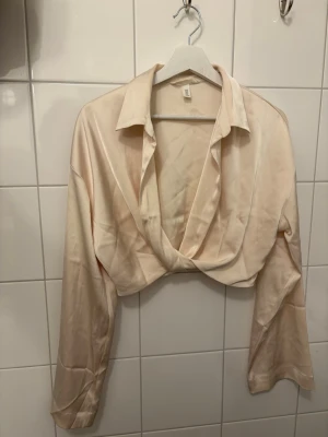 Krämvit omlottblus från H&M - Snygg krämvit omlottblus från H&M med djup v-ringning och klassisk krage. Blusen har långa ärmar och är tillverkad i ett glansigt, satinliknande material som ger en lyxig känsla. Perfekt att styla med jeans eller kjol för en trendig look.