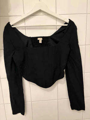 Svart långärmad korsettopp H&M - Snygg svart långärmad topp från H&M med korsettliknande form och bred, fyrkantig urringning. Toppen har smockad rygg för extra komfort och sitter tajt över kroppen. Perfekt att styla med jeans eller kjol för en trendig look.