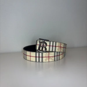 Rutigt bälte  - Säljer ett klassiskt bälte med det ikoniska beige, röda och svarta rutmönstret. Bältet har ett silvrigt B-spänne framtill och är gjort i läder med textil på ovansidan. Perfekt accessoar för att lyfta vilken outfit som helst.