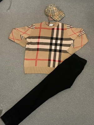 Beige rutig tröja från Burberry - Snygg beige tröja från Burberry med klassiskt rutmönster i svart, vitt och rött. Tröjan har rund halsringning, ribbade muddar och en avslappnad passform. Perfekt för dig som gillar tidlös och trendig stil.