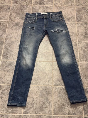 Replay anbass - Säljer ett par replay jeans med slitningar och hål på låren och vid fickorna. Jeansen har en klassisk femficksmodell och är tillverkade i denim med en tvättad look. Perfekta för en avslappnad och trendig stil.