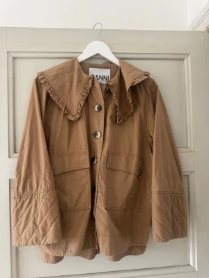 Beige jacka med volangkrage från Ganni - Trendig beige jacka från Ganni med stor volangkrage och dekorativa knappar framtill. Jackan har två stora fickor och quiltade detaljer på ärmarna. 