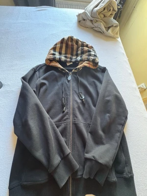 Svart hoodie från Burberry med mönstrad huva - Svart hoodie från Burberry med dragkedja och klassisk rutmönstrad huva i beige, svart och rött. Hoodien har ribbade muddar, fickor framtill och justerbara snören vid huvan. Perfekt för en stilren och lyxig streetwear-look. Passar M perfekt 