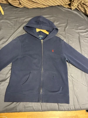 Marinblå hoodie från Polo Ralph Lauren - Klassisk marinblå hoodie från Polo Ralph Lauren med dragkedja framtill och kängurufickor. Ikoniska röda Polo-loggan broderad på bröstet. Tillverkad i mjuk bomullsblandning och har en stor huva för extra chill vibe.