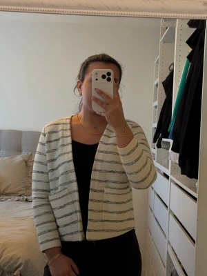 Vit kofta med svarta ränder - Säljer en stilren vit kofta med tunna svarta horisontella ränder. Koftan har öppen front, två fickor framtill och långa ärmar. Perfekt att slänga över en topp för en enkel men snygg look.