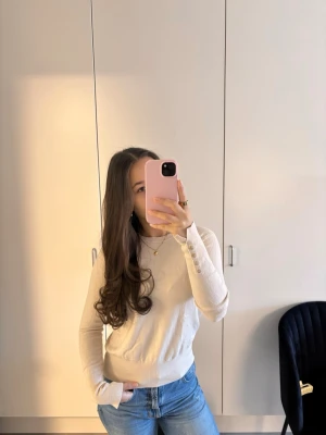 Beige stickad tröja med gulddetaljer - Snygg beige stickad tröja med rund hals och långa ärmar. Tröjan har ribbade muddar och dekorativa guldfärgade knappar vid ärmsluten som ger en exklusiv touch. Perfekt att matcha med jeans för en stilren look.