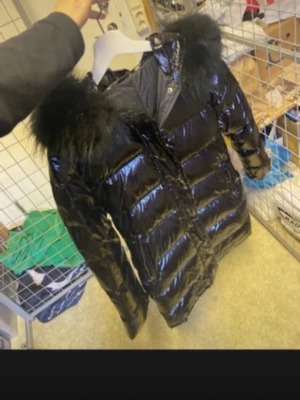 Glansig svart dunjacka med päls - Använd en gång Snygg svart dunjacka med glansigt material och stor huva med svart päls Jackan är lång, har quiltad design och stängs med dragkedja och knappar. Perfekt för kalla vinterdagar och ger en riktigt trendig look.