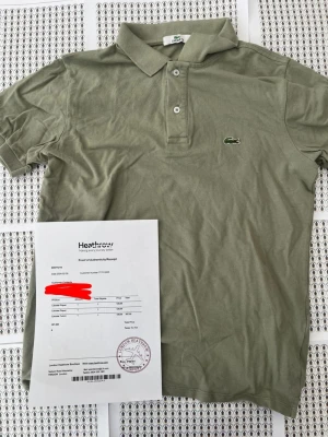 Olivgrön pikétröja från Lacoste - Säljer en klassisk olivgrön pikétröja från Lacoste med den ikoniska krokodilloggan på bröstet. Tröjan har korta ärmar, krage och knäppning med två knappar framtill. Perfekt till jeans eller shorts för en clean och stilren look.