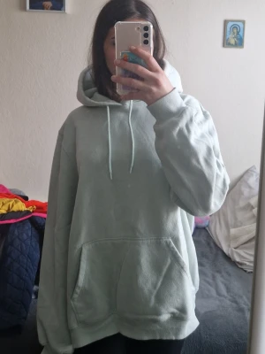 Ljusgrön oversized hoodie - Mysig ljusgrön hoodie med huva och dragsnören. Modellen är oversized med stor känguruficka framtill och ribbade muddar vid ärmar och nederkant. Perfekt för en chill och avslappnad stil. Materialet känns mjukt och bekvämt, troligen bomullsblandning.