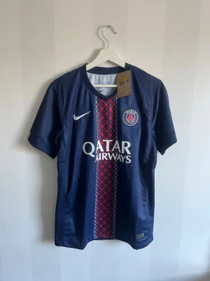 PSG matchtröja O. Dembélé nr 10 - Säljer en mörkblå PSG matchtröja från Nike med O. Dembélé och nummer 10 på ryggen. Tröjan har korta ärmar, röd och vit randig detalj framtill samt Qatar Airways-logga. Perfekt för fotbollsfans som vill stötta sitt lag.