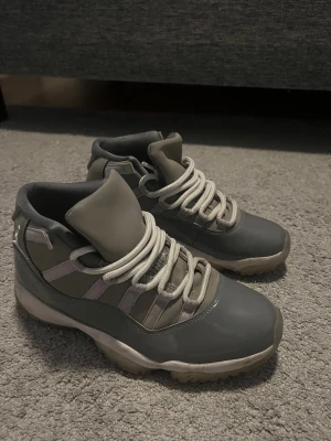 Gråa Air Jordan 11 sneakers - Snygga Air Jordan 11 sneakers i olika nyanser av grått med vita skosnören. Skorna har en blank tå i syntetmaterial och meshpaneler på sidorna. Klassisk high-top design som ger extra stöd runt ankeln. Perfekta för dig som gillar streetwear och sportig stil.