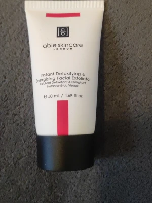 Able Skincare ansiktsskrubb 50 ml - Instant Detoxifying & Energising Facial Exfoliator från Able Skincare London. Vit tub med svart lock och rosa detalj, innehåller 50 ml. Perfekt för dig som vill ha en uppfriskande och rengörande ansiktsskrubb.