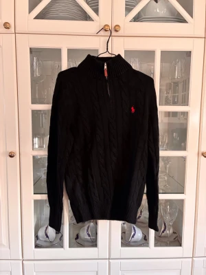 Svart kabelstickad tröja Polo Ralph Lauren - Svart kabelstickad tröja från Polo Ralph Lauren med halv dragkedja och hög krage. Klassisk röd logga broderad på bröstet. Tröjan har ribbade muddar och är perfekt för lager-på-lager-stil.