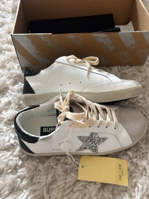 Golden Goose - Snygga vita Golden Goose sneakers med silverglittrig stjärna på sidan, svart glitterdetalj på hälen och beige mockadetalj vid tån. Skorna har vita snören och klassisk låg modell. Perfekta sommarskor! Säljer pga för små!