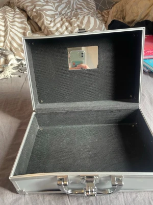 Silvrig sminkväska med spegel - Praktisk sminkväska i silverfärgad metall med robust handtag och lås. Insidan är klädd i svart tyg och locket har en liten spegel. Perfekt för att förvara smink, borstar eller andra skönhetsverktyg. Stilren och enkel design som passar alla.