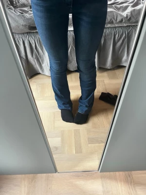 Blå bootcut jeans med medelmidja - Säljer ett par klassiska blå bootcut jeans med medelhög midja och smal passform upptill som går ut något vid bensluten. Jeansen har fem fickor och stängs med dragkedja och knapp. Perfekta till sneakers eller boots! Obs! Se bild 5 att det är ett litet slitage på ena benet.