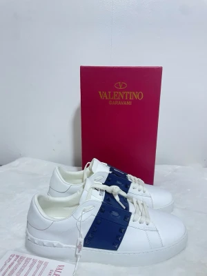 Valentino Garavani Rockstud sneakers - Snygga vita sneakers från Valentino Garavani med en bred mörkblå rem över snörningen dekorerad med ikoniska nitar. De är helt nya och i bästa skick du kan hitta, kvitto skickas med. Hör av dig om du vill ha mer info🥂. Skicka prisförslag vid snabb affär!                    Skick[10/10]. Storlek[43]. Pris[3599]