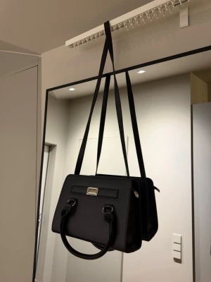 Svart handväska med axelrem - Svart handväska i fyrkantig modell med handtag och lång axelrem. Aldrig använd.