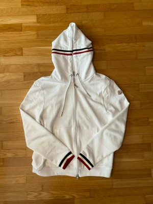 Vit Moncler hoodie med ränder - Snygg vit hoodie från Moncler med dragkedja framtill och huva. Har röda och marinblå ränder på huvan och vid ärmsluten samt Moncler-logga på ena ärmen. Tillverkad i mjukt bomullsmaterial och har fickor framtill. Perfekt för en clean och sportig look. Säljer min vita Moncler zip eftersom den är för stor för mig. Dm mig för fler frågor.